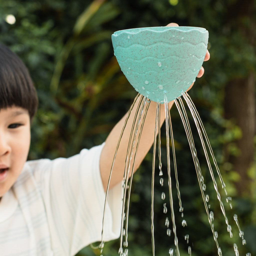 Young child pouring water from Bath Egg stack outside, watching water stream through drainage holes, engaging in cause-and-effect learning.
