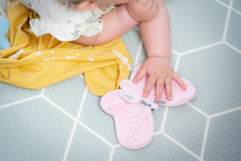 Close-up of baby sitting on a tiled floor grasping the pink butterfly scrunchie toy, illustrating gentle sensory interaction and fine motor exploration.
