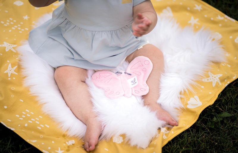 Baby sitting on a yellow blanket on grass, playing with the pink butterfly scrunchie toy, enjoying tactile play with its crinkle wings and soft fabric.