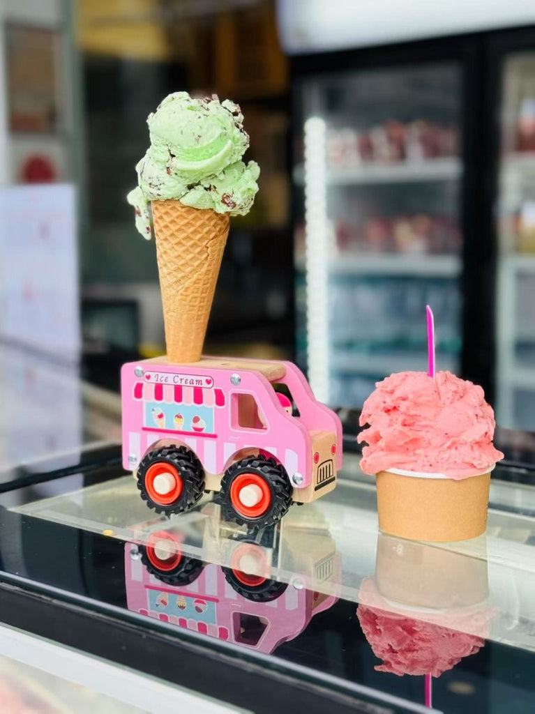 Pink toy truck with a green ice cream cone on top, next to a pink ice cream cone with a pink toy car on a reflective surface.