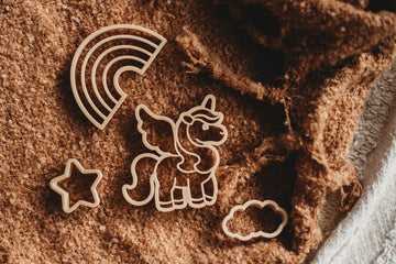 Flat lay of Kinfolk Pantry Unicorn Cutter Set on brown fabric, featuring unicorn, rainbow, cloud, and star shapes made from eco-friendly materials.