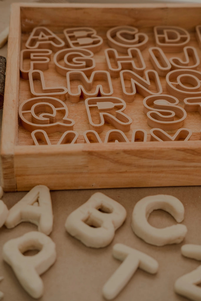 Eco plastic alphabet cutters with letters A to Z on a wooden tray, surrounded by dough letters.