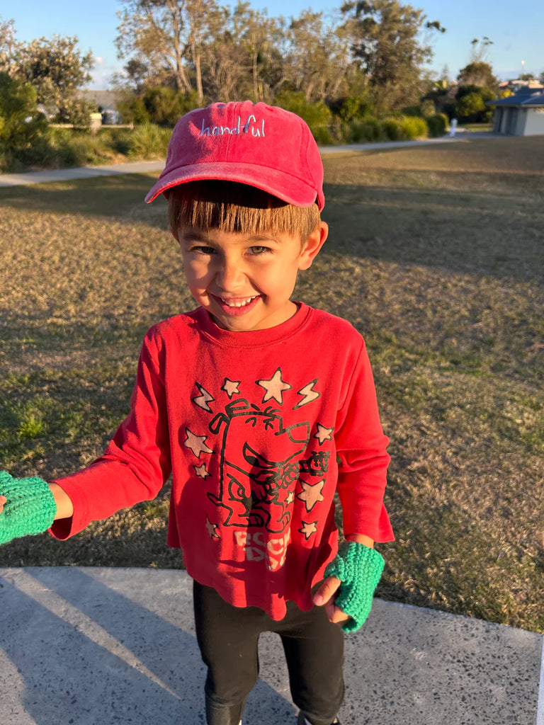 Young child wearing the watermelon red "Handful" cap with a matching red outfit, standing outdoors—showcasing the bright, bold style of the cap in action.