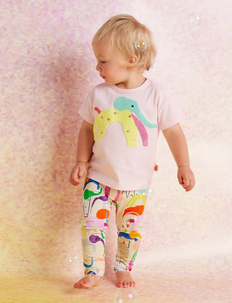 Toddler in Halcyon Nights Happy Herd Baby Leggings and matching pink t-shirt, smiling and walking barefoot against a soft pink textured backdrop.