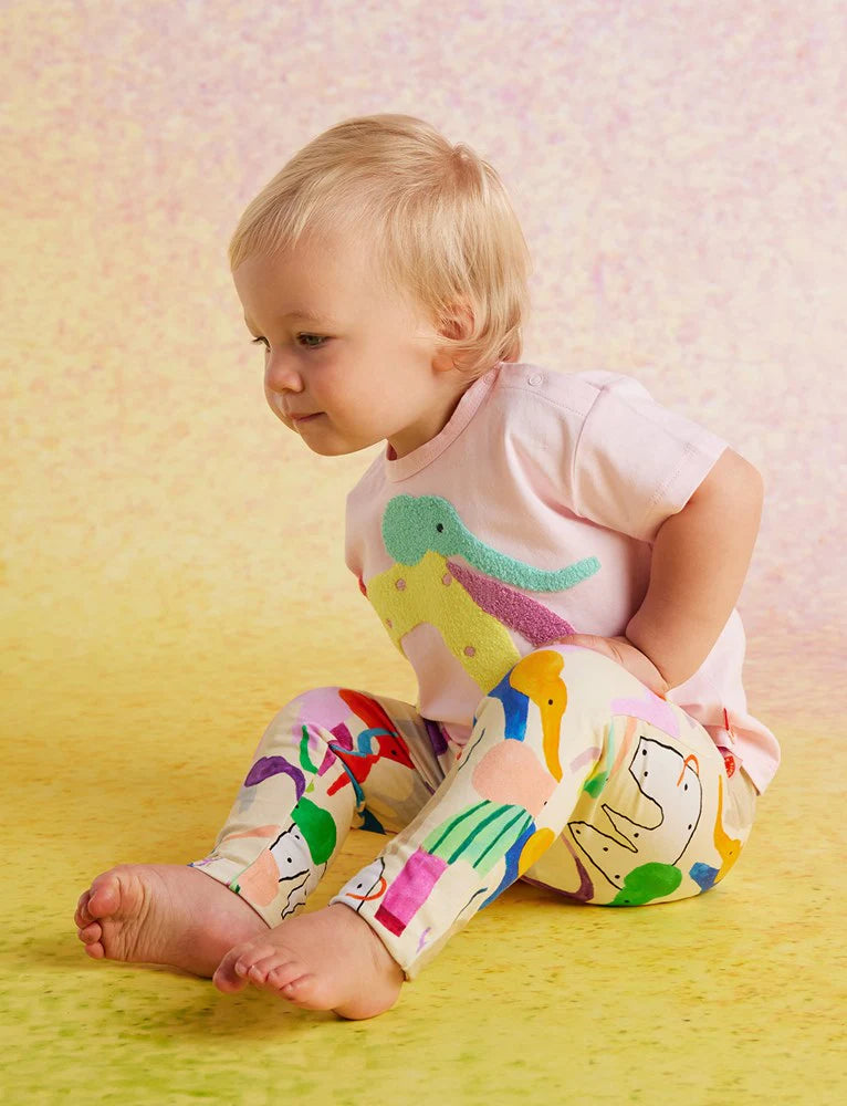 Toddler wearing Halcyon Nights Happy Herd Baby Leggings and a light pink elephant t-shirt, seated playfully on a yellow pastel background.