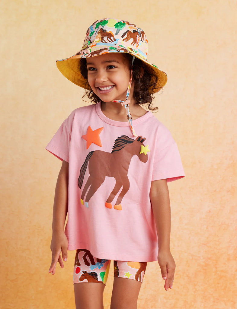 Child in Halcyon Nights Giddy Up Bike Shorts and pink horse tee standing confidently with hands relaxed and a cheerful expression.