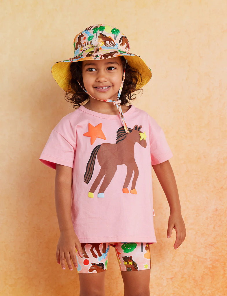 Smiling child wearing Halcyon Nights Giddy Up Bike Shorts and matching pink horse tee with a wide-brimmed hat, standing against a peach background.