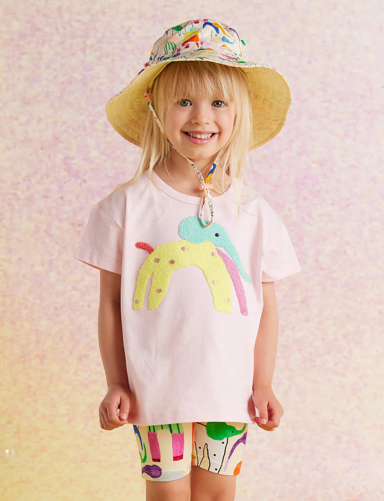 Young girl wearing Happy Herd Bike Shorts with a white printed t-shirt and wide-brimmed floral hat, standing playfully against a pink gradient background.