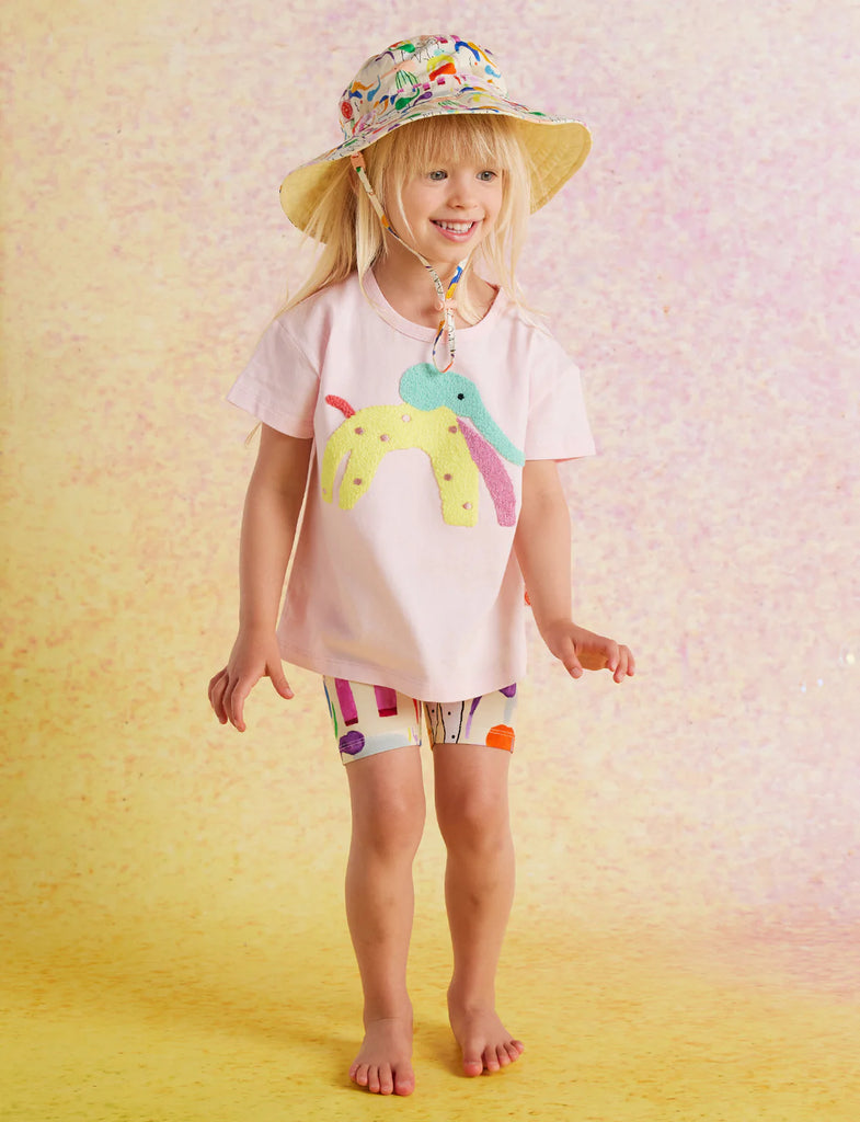 Close-up of girl smiling in Happy Herd Bike Shorts and a white graphic t-shirt, styled with a summery hat, posing against a soft yellow-pink background.