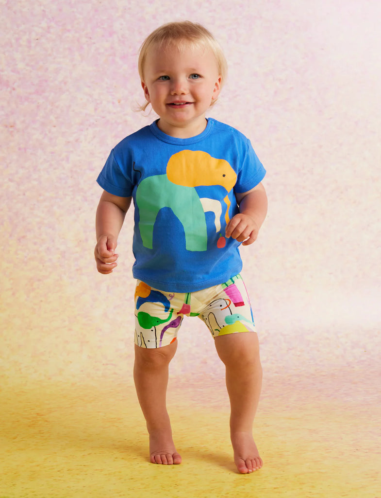 Smiling toddler wearing Halcyon Nights Happy Herd Bike Shorts and a bright blue t-shirt, standing barefoot on a yellow background.