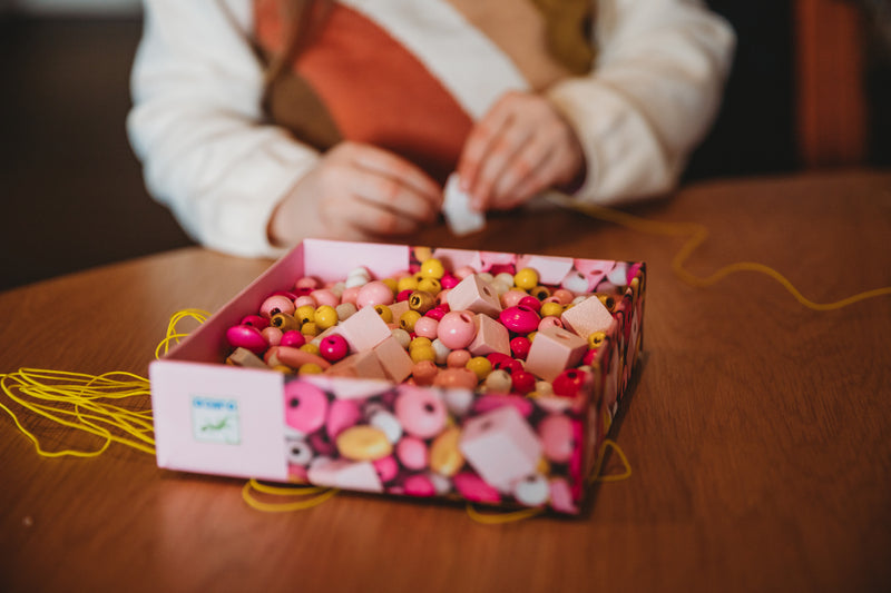 Close-up of child’s hands reaching into the open Djeco bead box, selecting beads for threading activity.