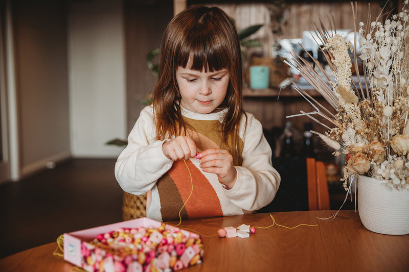 Child threading pink wooden beads from Djeco bird-themed set at a wooden table, focusing intently on the crafting activity.