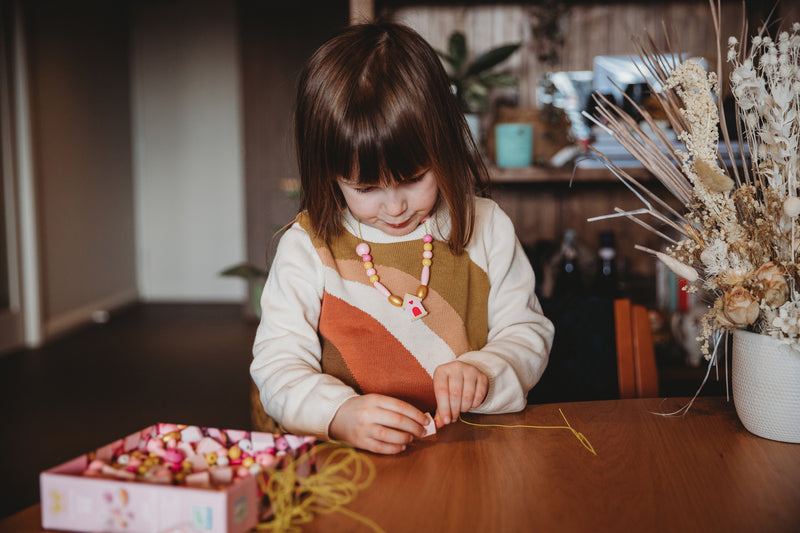 Young girl seated at a table wearing a beaded necklace she made using Djeco bird-themed beads, with packaging in front of her.
