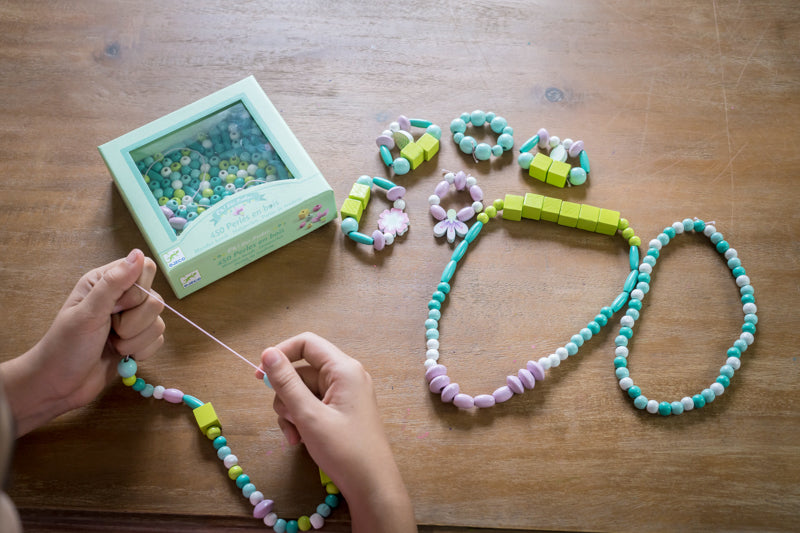 Child making beaded necklaces with the full Djeco bead set spread out, including colourful beads and flower charms on a wooden surface.