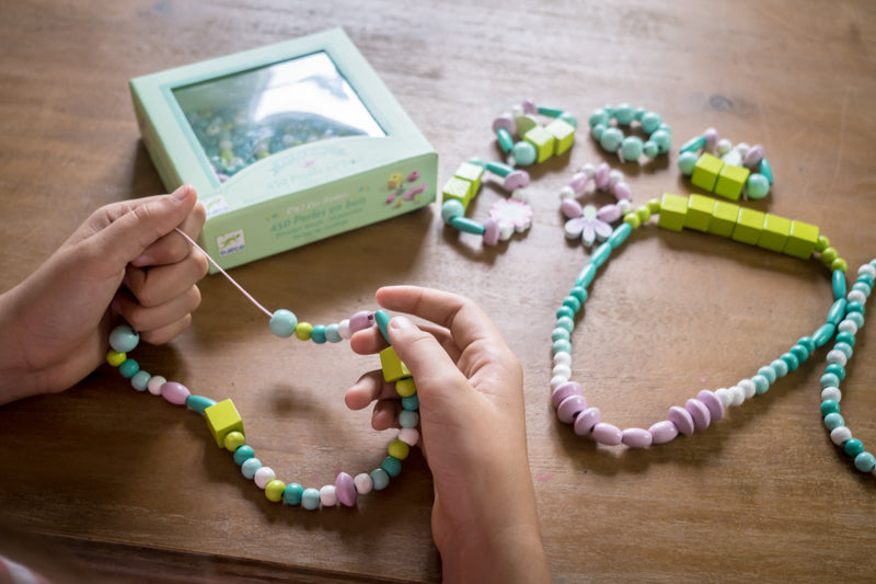 Child creating beaded jewellery using Djeco wooden beads on a table, with completed pieces and the packaging box nearby.