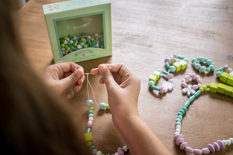Close-up of child’s hands threading pastel wooden beads onto string, with Djeco bead set box visible in the background.