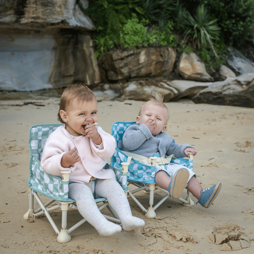 Two babies sitting on a sandy beach in Izimini Baby Chairs with blue and green checkered designs, smiling and eating snacks.