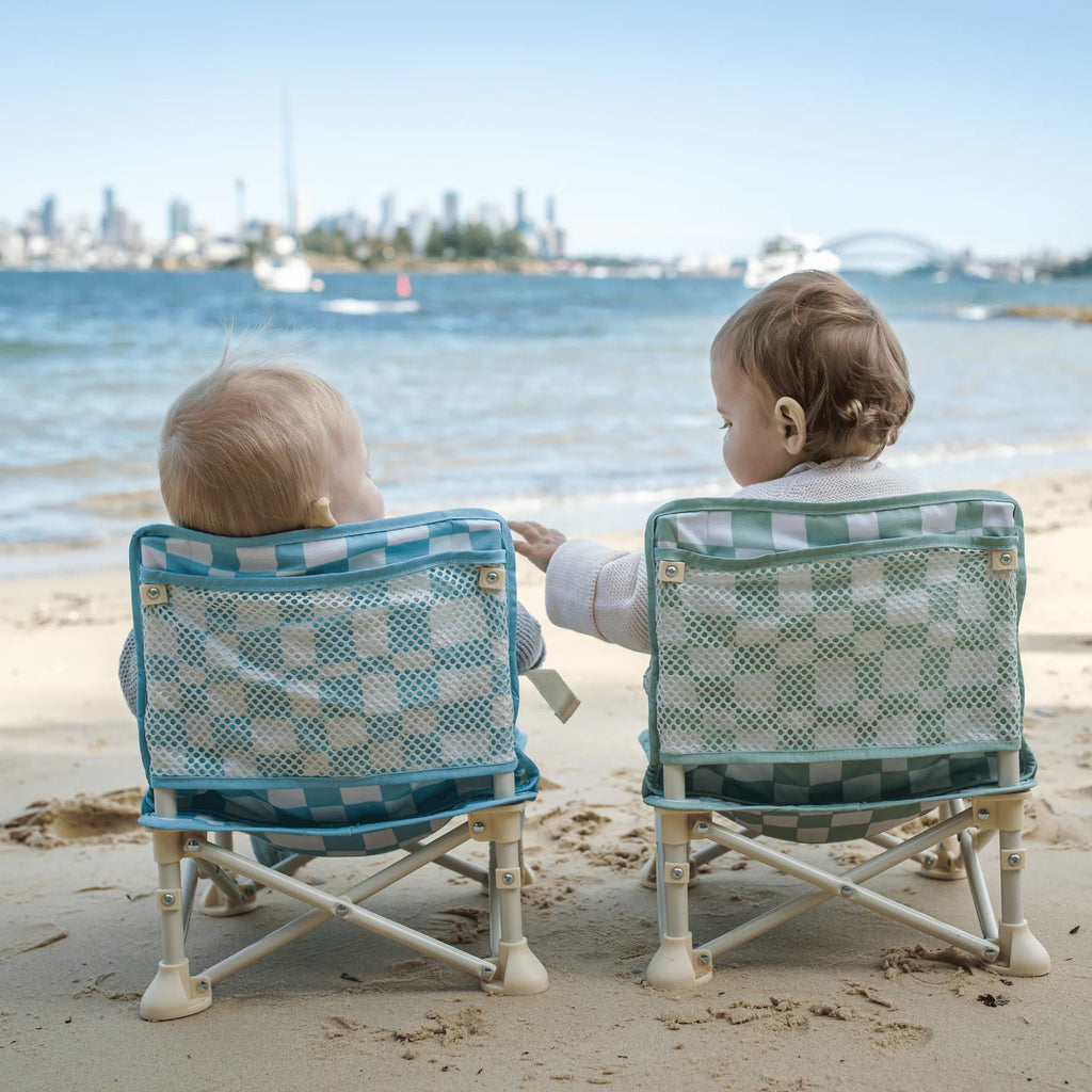 Rear view of two babies sitting side by side in Izimini Baby Chairs with blue and green checkered patterns, overlooking the ocean and city skyline.