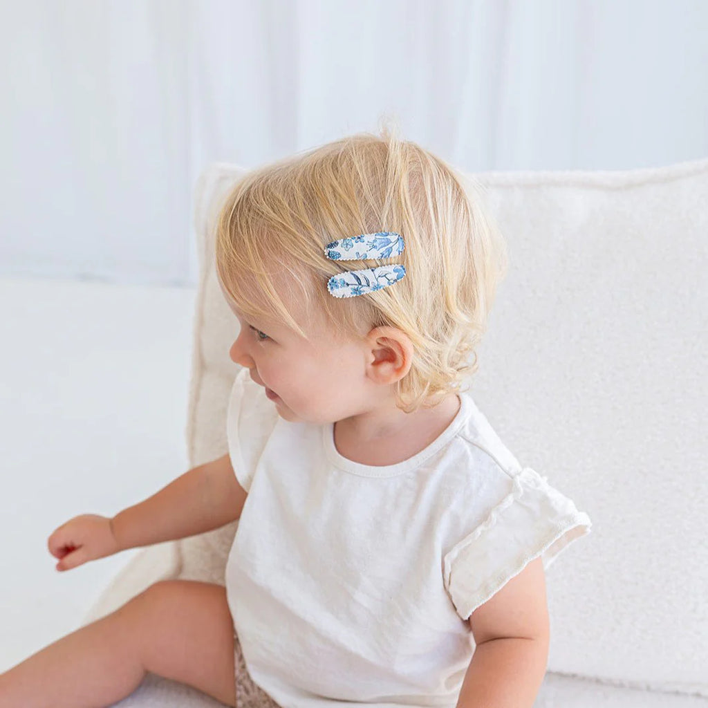 Child with blonde hair wearing two blue hair clips sitting on a white couch.