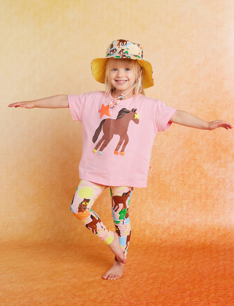 Smiling child in Giddy Up Kids Leggings, pink horse tee, and bucket hat, standing with arms outstretched against an ombré orange background.