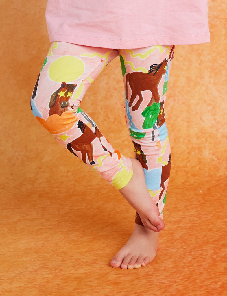 Child wearing Halcyon Nights Giddy Up Kids Leggings with visible horse print, standing on one foot against an orange backdrop.