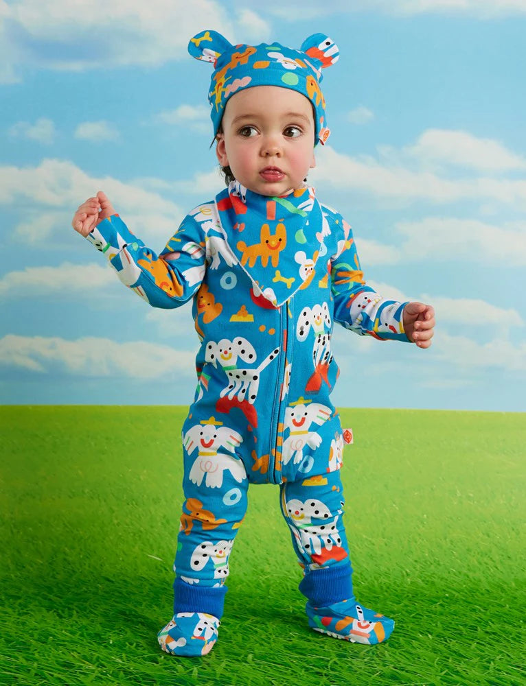 Front view of baby standing in the Halcyon Nights Puppy Park long sleeve zip romper, showcasing bold, colourful puppy and sunshine print.