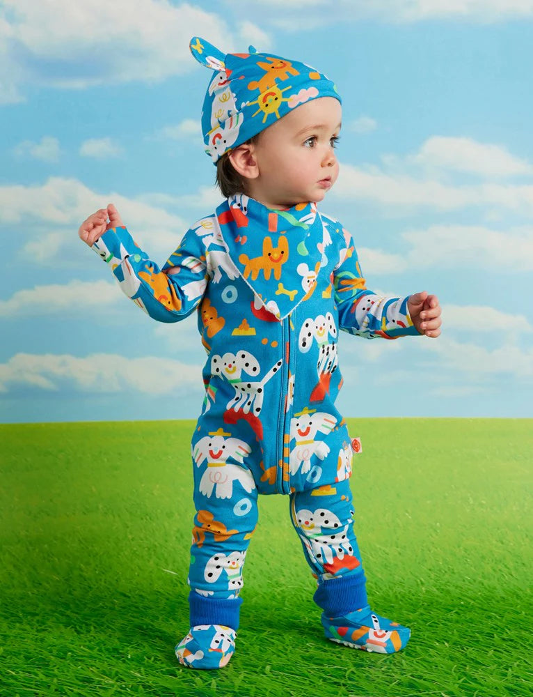 Baby wearing the Halcyon Nights Puppy Park romper and matching hat, standing on bright green grass with a blue sky background.