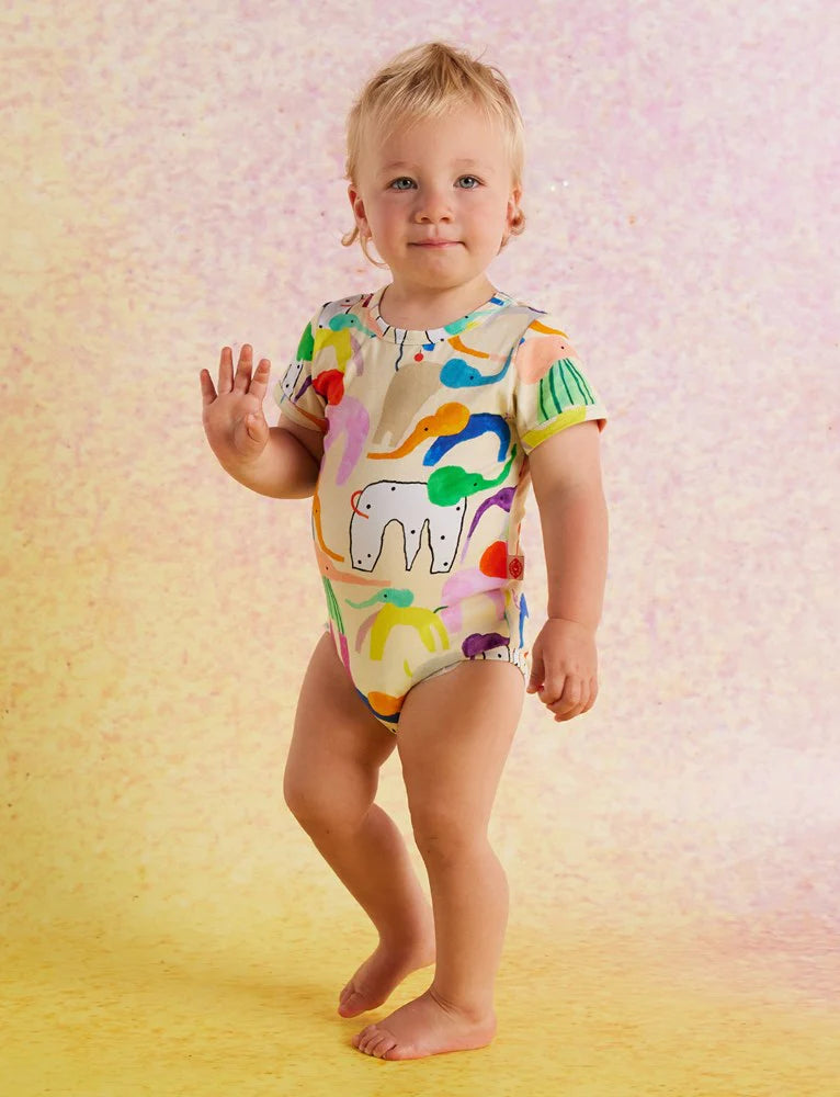 Toddler standing and smiling in the Happy Herd short sleeve bodysuit by Halcyon Nights, showing off the playful and colourful animal print.