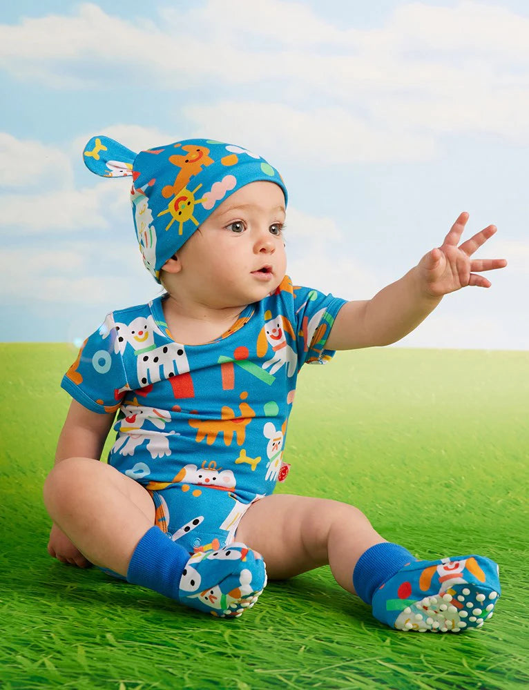 Baby wearing the Puppy Park short sleeve bodysuit and matching hat, sitting on green grass with a blue sky background, reaching with one hand.