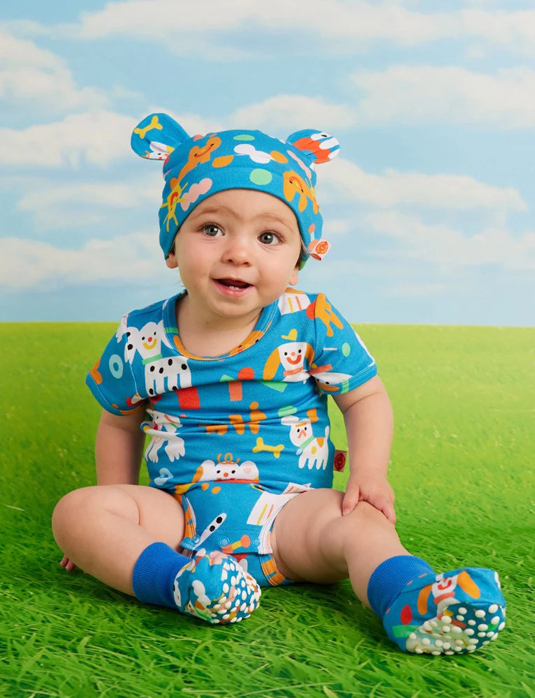 Smiling baby seated in the Halcyon Nights Puppy Park short sleeve bodysuit and matching accessories, posed on green grass against a sky-themed backdrop.
